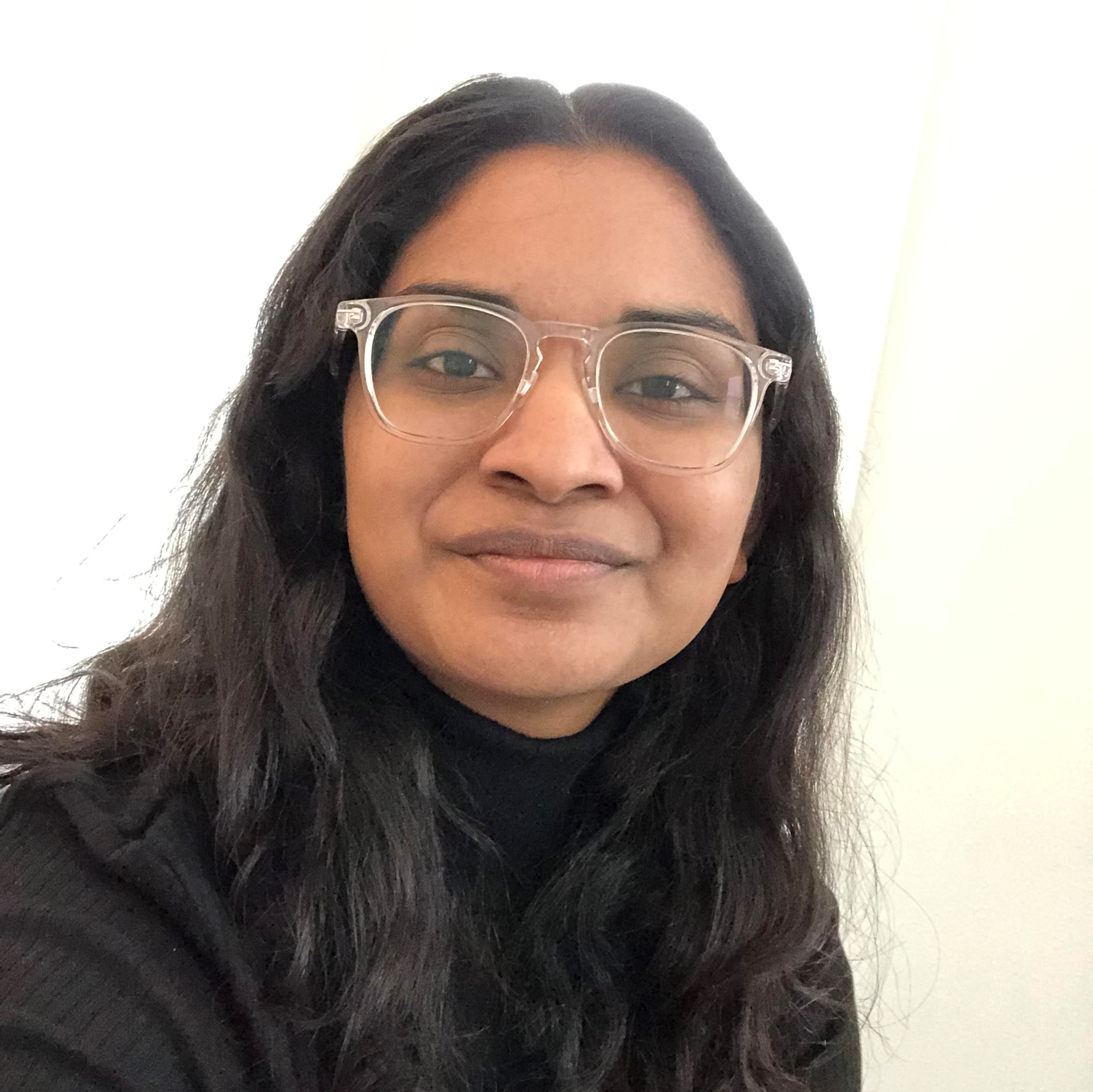 A picture of me (Sruthi) smiling, wearing a black turtleneck and clear glasses!